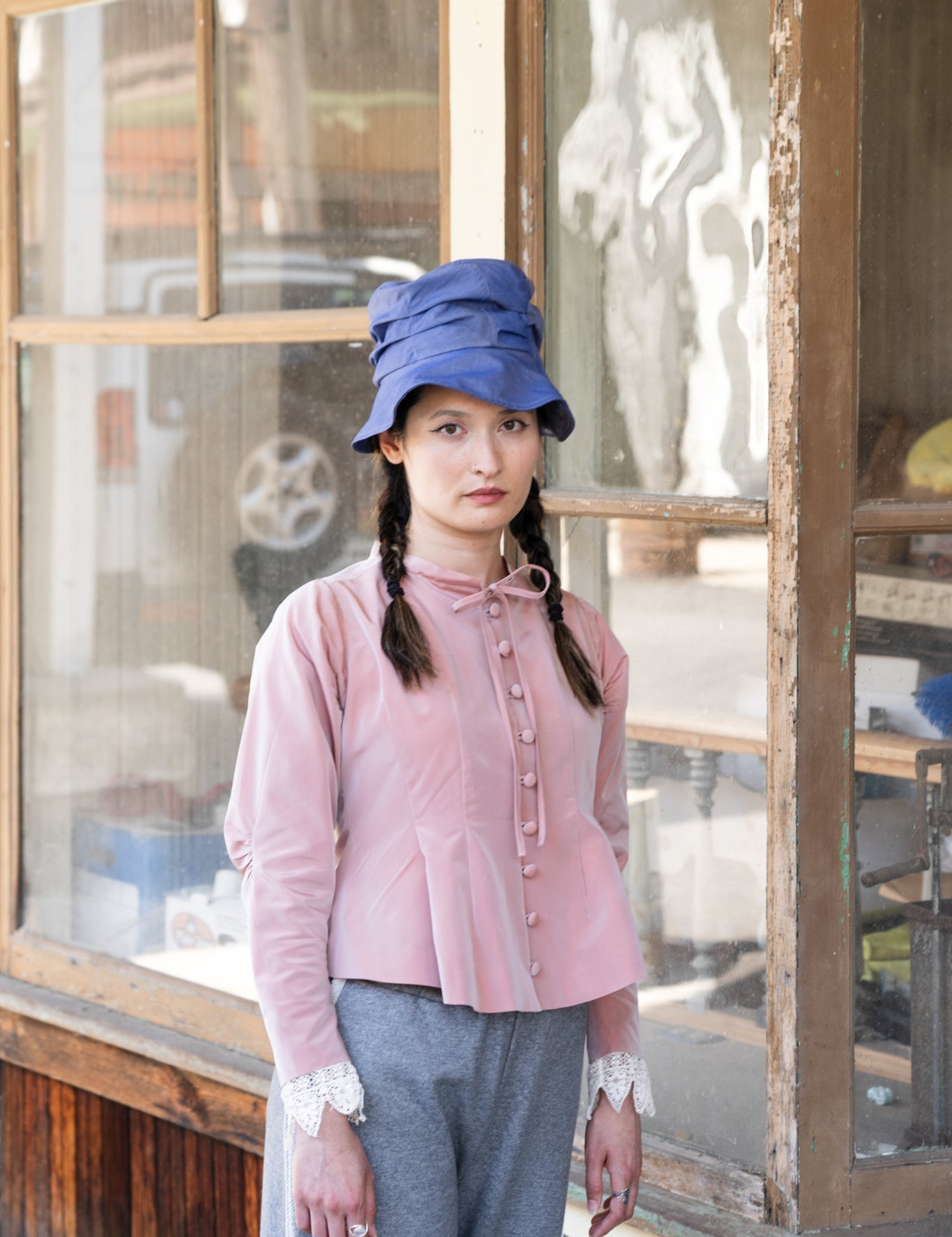 Woman wearing a pink blouse and blue hat in front of a wooden window.