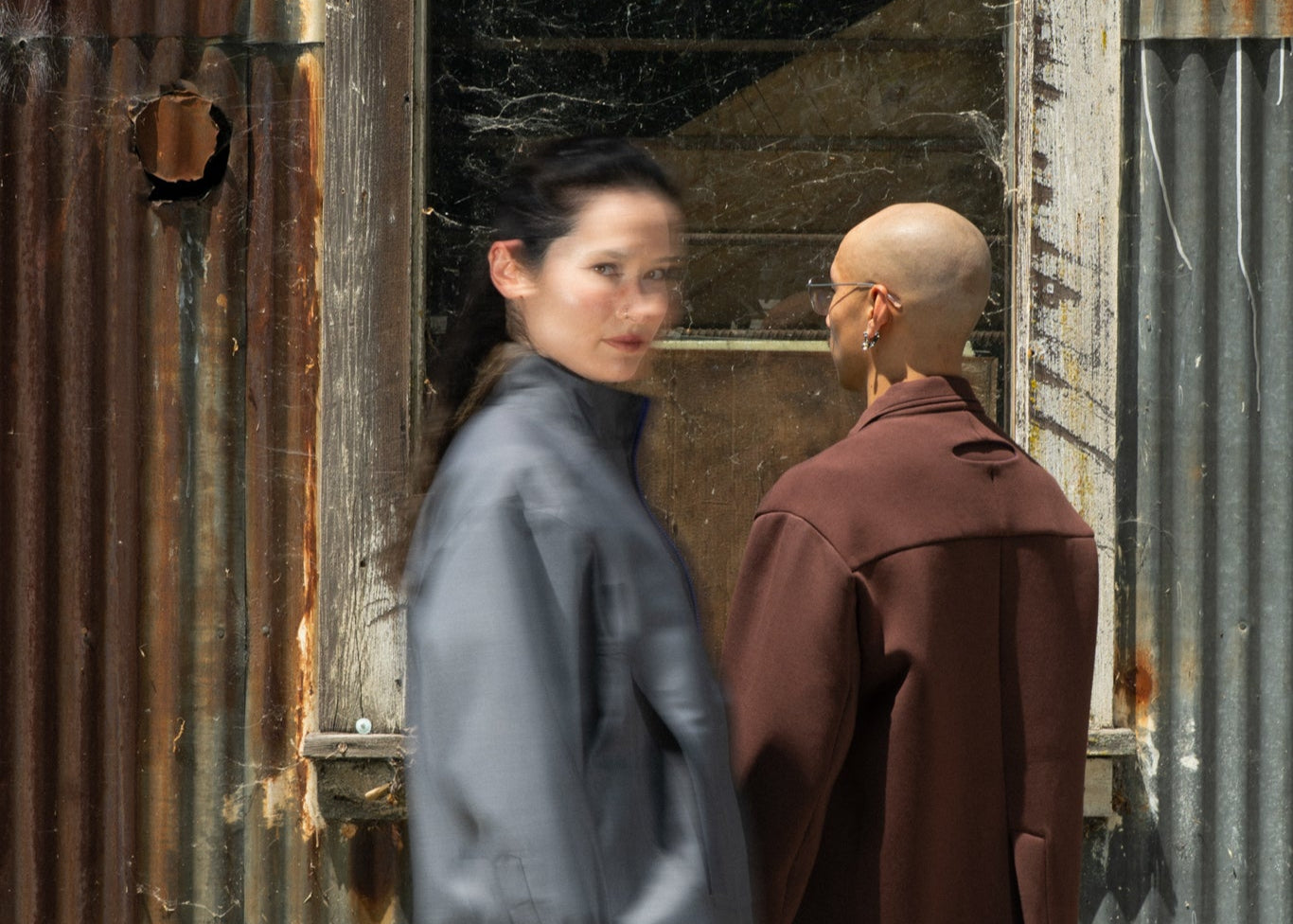Two people standing in front of a rusty corrugated metal wall.
