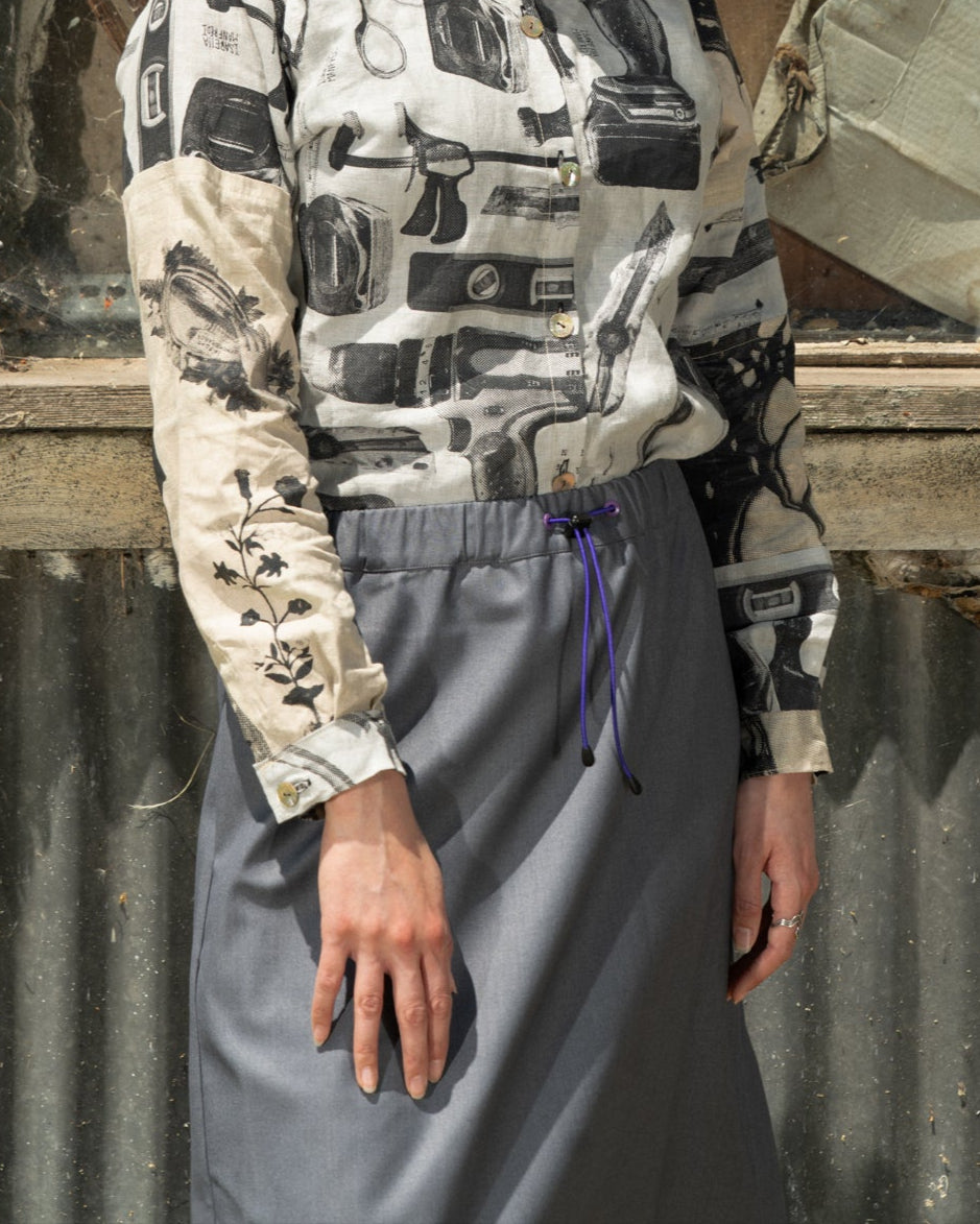 Woman in a patterned blouse and gray skirt standing in front of a reflective surface.