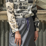 Woman in a patterned blouse and gray skirt standing in front of a reflective surface.