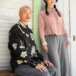 Two people posing outdoors, one sitting on a bench and the other standing against a wooden wall.