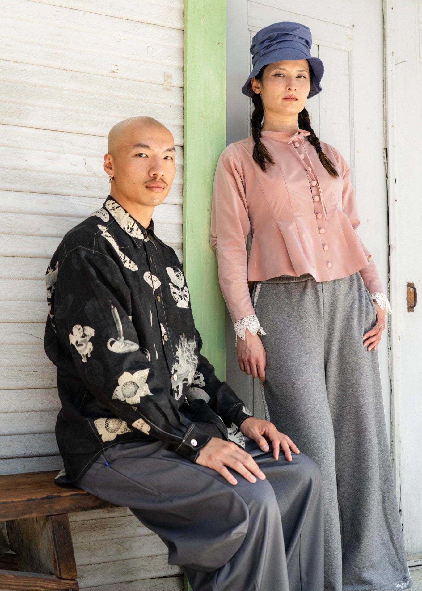 Two people posing outdoors, one sitting on a bench and the other standing against a wooden wall.