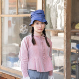 Woman wearing a pink blouse and blue hat in front of a wooden window.