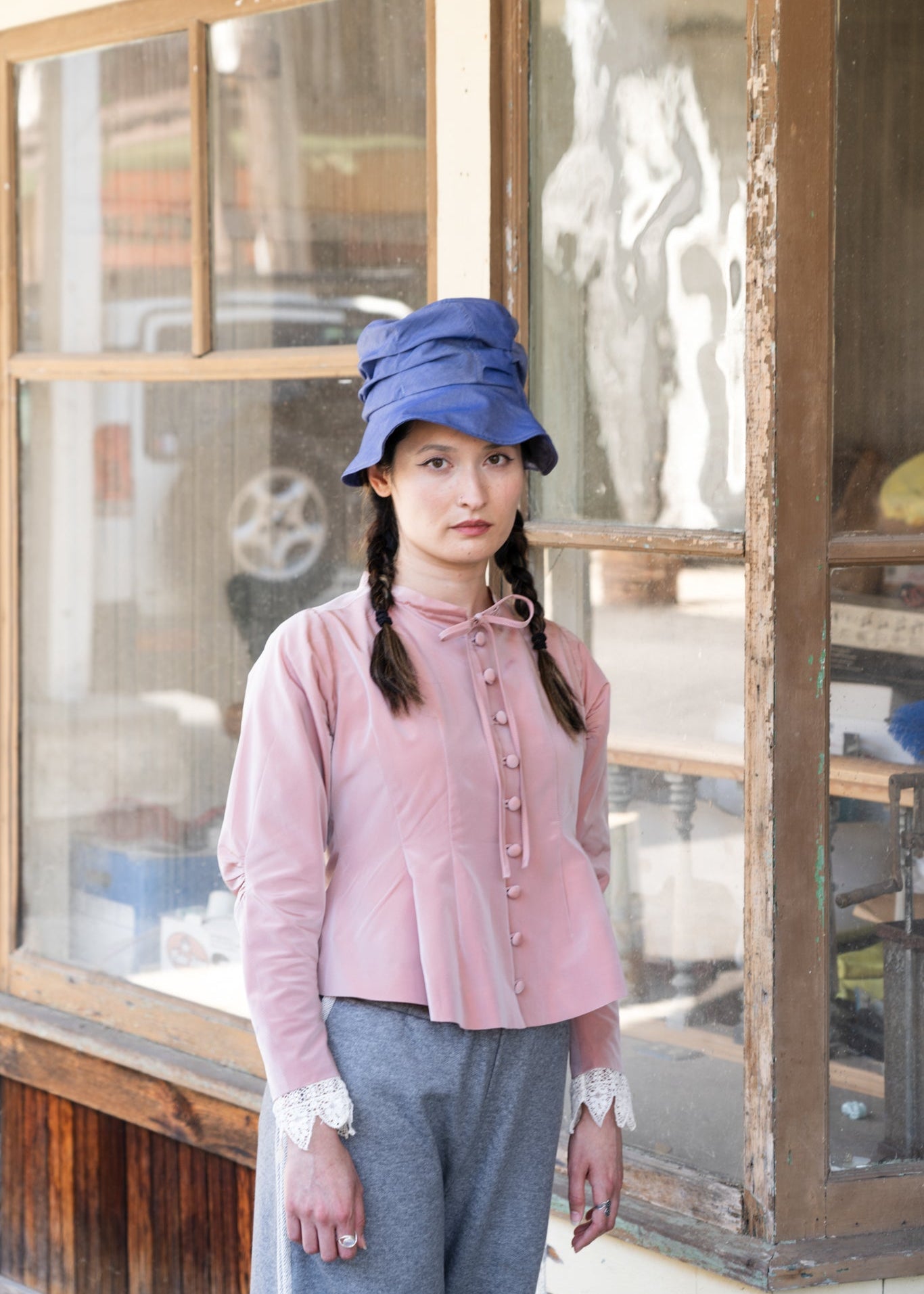 Woman wearing a pink blouse and blue hat in front of a wooden window.
