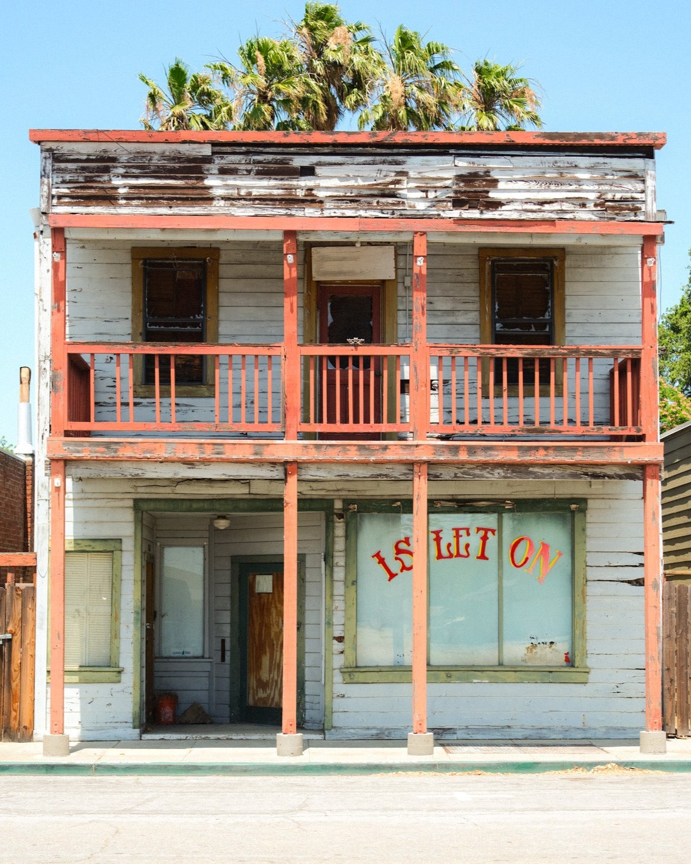 Old two-story building with a balcony and 'For Rent' sign on a sunny day.